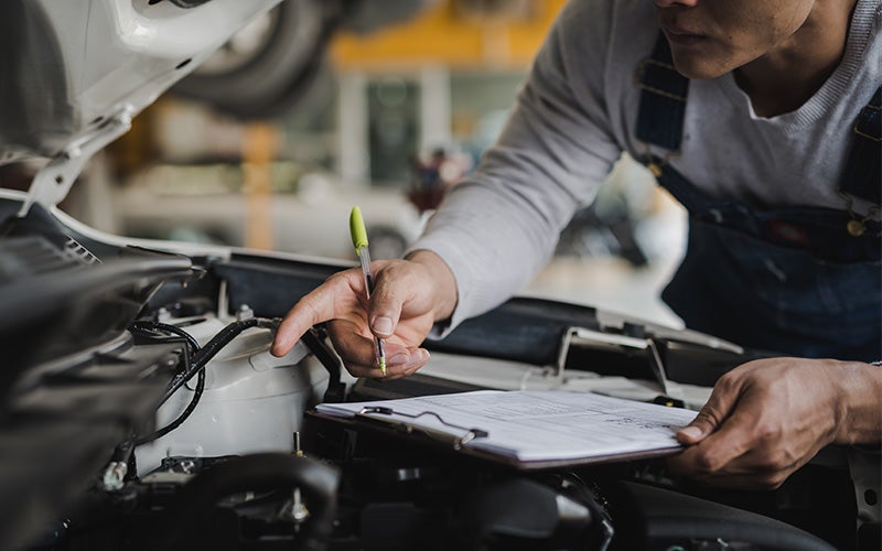 Mechanic in overalls pointing at a car engine while holding a clipboard and pen.